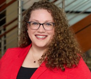 Headshot of Suzy Finn, a Caucasian woman with brown curly hair, wearing glasses and a red jacket over a black shirt